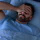Man with a beard lies in bed under blue bedding, hand covering his eyes in a gesture of fatigue or stress.