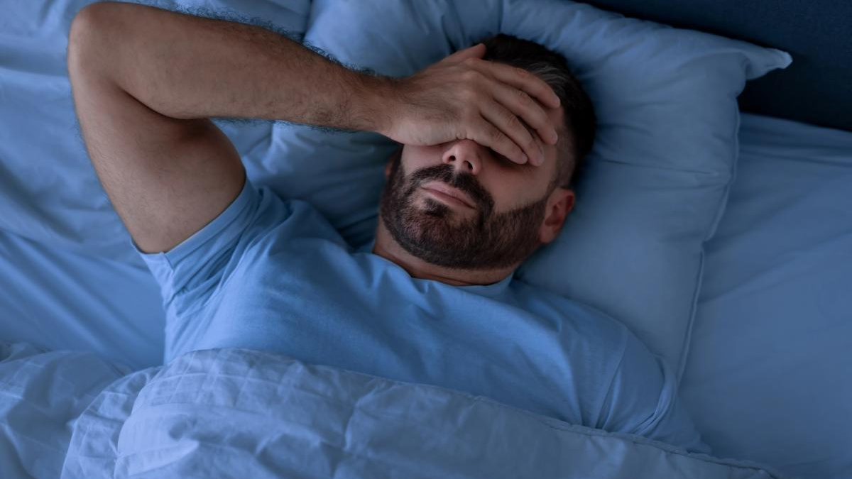 Man with a beard lies in bed under blue bedding, hand covering his eyes in a gesture of fatigue or stress.