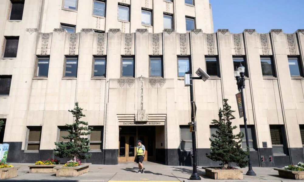 Front entrance of a beige art-deco building labeled 'Tacoma Building' with a man in a safety vest walking by, on a city street.
