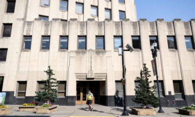 Front entrance of a beige art-deco building labeled 'Tacoma Building' with a man in a safety vest walking by, on a city street.