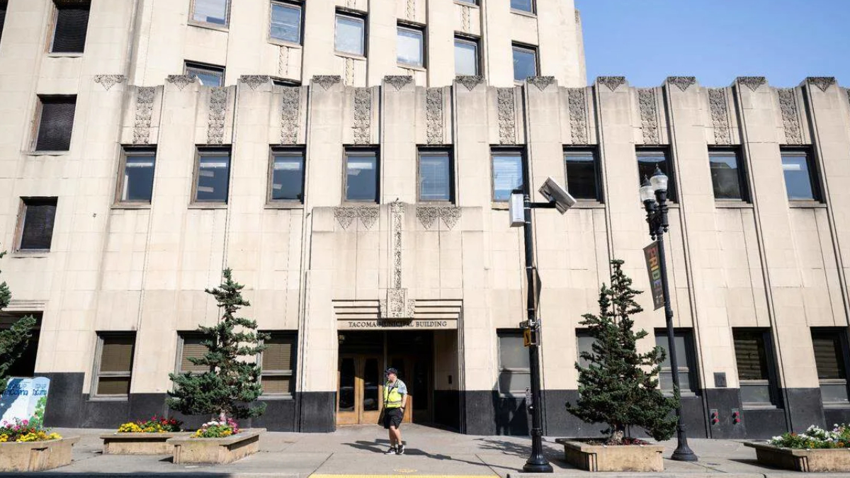 Front entrance of a beige art-deco building labeled 'Tacoma Building' with a man in a safety vest walking by, on a city street.