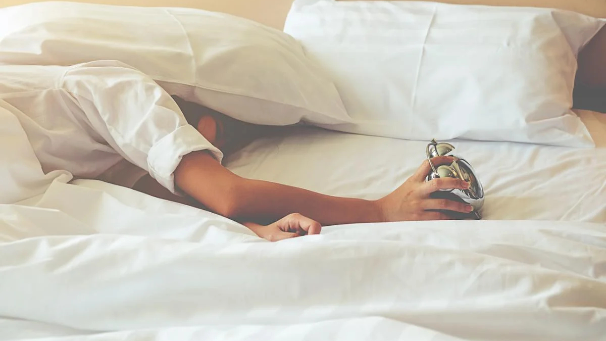 Person partially buried under white sheets reaching for a small silver alarm clock on a bed