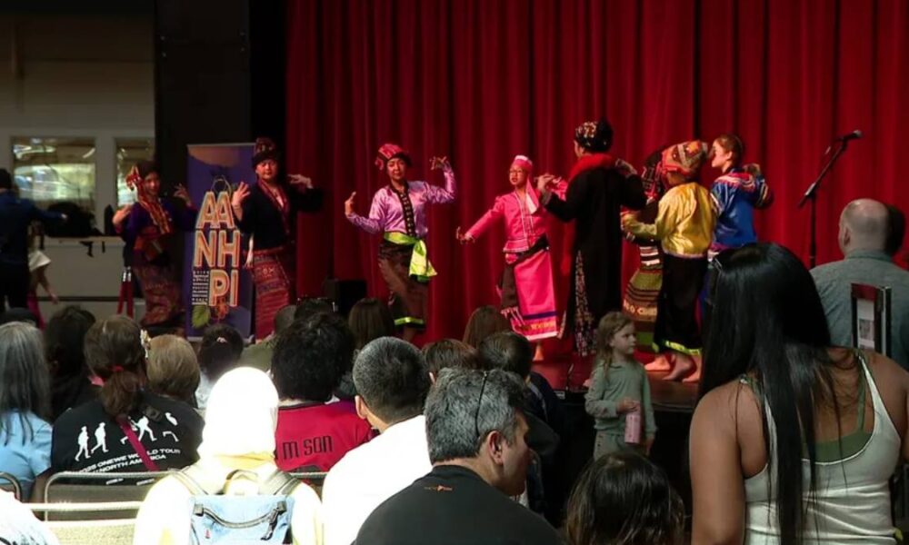A group of performers in colorful traditional costumes dancing on a stage with a red curtain, audience seated and watching.