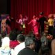 A group of performers in colorful traditional costumes dancing on a stage with a red curtain, audience seated and watching.