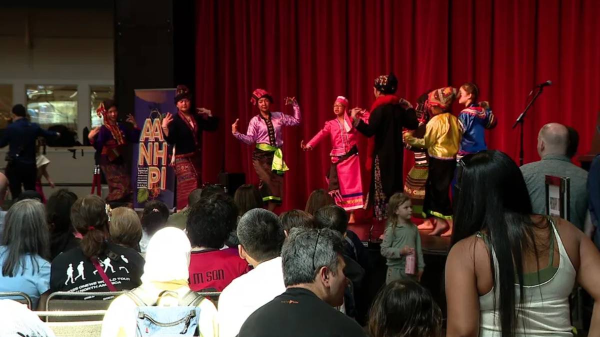 A group of performers in colorful traditional costumes dancing on a stage with a red curtain, audience seated and watching.