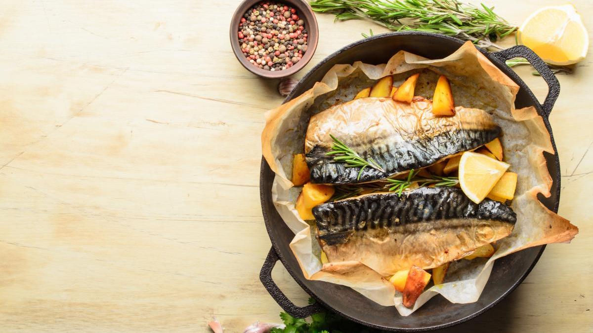 Two roasted mackerel fillets with potato chunks and lemon wedges in a parchment-lined cast-iron skillet, garnished with rosemary.