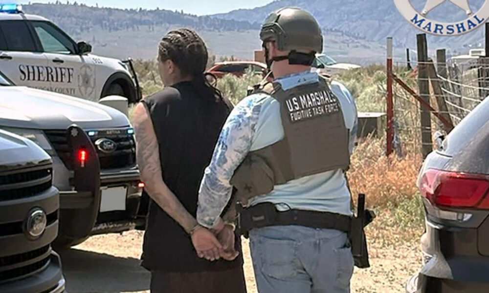 Suspect in handcuffs escorted by a U.S. Marshal wearing a tactical vest, with sheriff and patrol vehicles in a rural setting behind them.