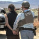 Suspect in handcuffs escorted by a U.S. Marshal wearing a tactical vest, with sheriff and patrol vehicles in a rural setting behind them.