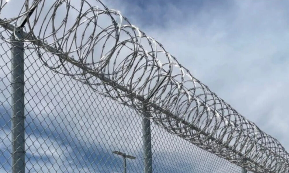 Chain-link fence topped with spiraled razor wire against a cloudy blue sky