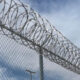 Chain-link fence topped with spiraled razor wire against a cloudy blue sky