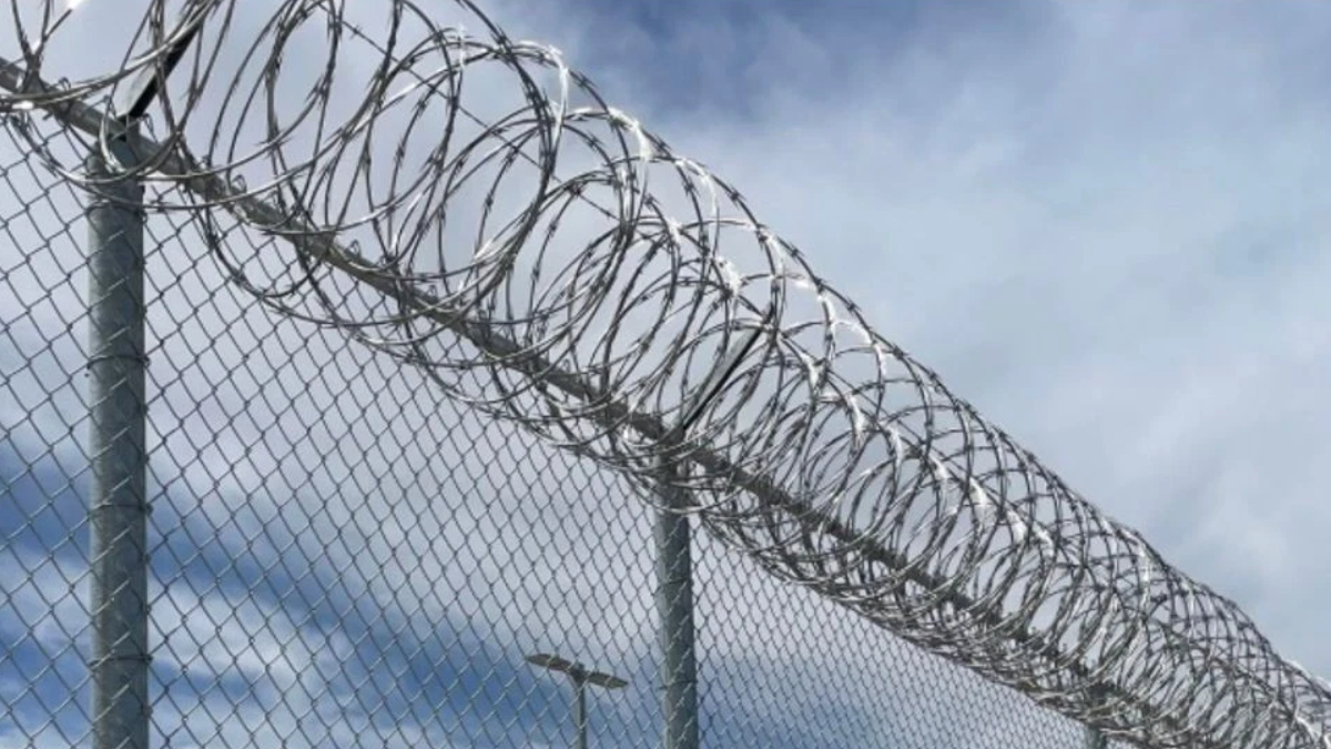 Chain-link fence topped with spiraled razor wire against a cloudy blue sky