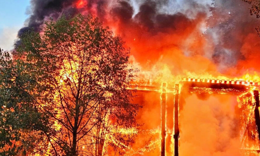Wooden frame building engulfed in bright flames with thick smoke rising behind trees in the foreground.