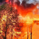 Wooden frame building engulfed in bright flames with thick smoke rising behind trees in the foreground.