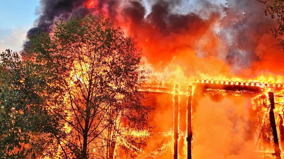 Wooden frame building engulfed in bright flames with thick smoke rising behind trees in the foreground.