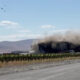 Building on fire along a rural highway with thick black smoke and emergency vehicles on scene near a vineyard fence.