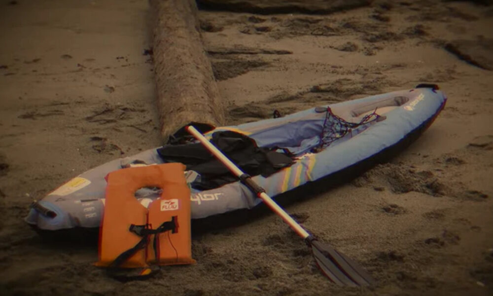 Inflatable kayak with a paddle and orange life jacket placed on a sandy beach, ready for paddling.