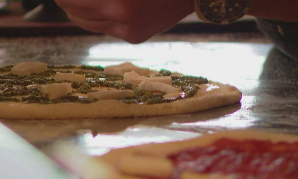Pizza dough base with pesto and cheese toppings being added by hand on a work surface