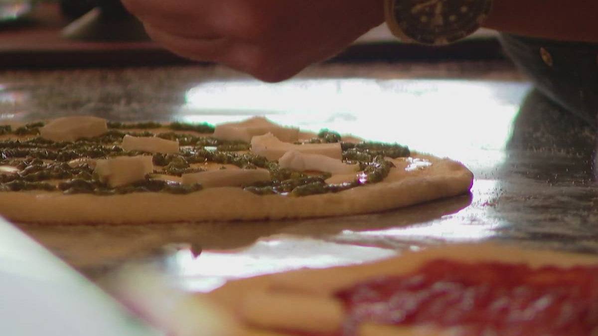 Pizza dough base with pesto and cheese toppings being added by hand on a work surface