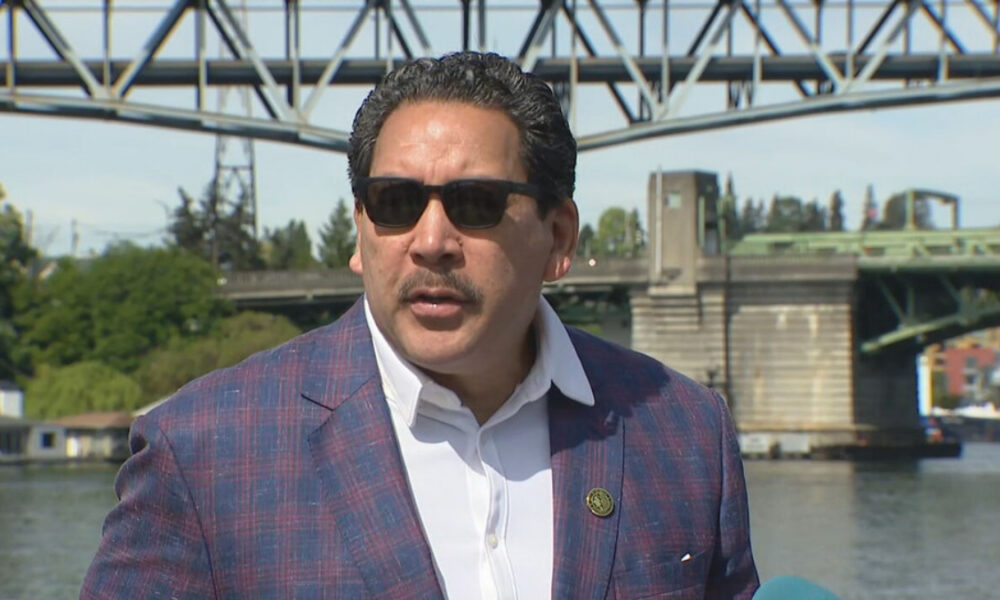 Man in a blue plaid blazer and white shirt wearing sunglasses speaks during a news interview by a river with a metal bridge in the background