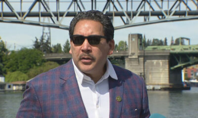 Man in a blue plaid blazer and white shirt wearing sunglasses speaks during a news interview by a river with a metal bridge in the background