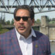 Man in a blue plaid blazer and white shirt wearing sunglasses speaks during a news interview by a river with a metal bridge in the background