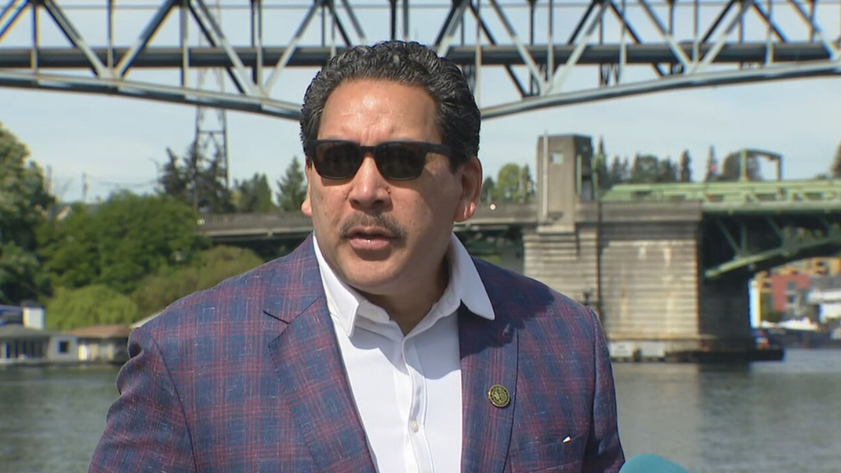 Man in a blue plaid blazer and white shirt wearing sunglasses speaks during a news interview by a river with a metal bridge in the background