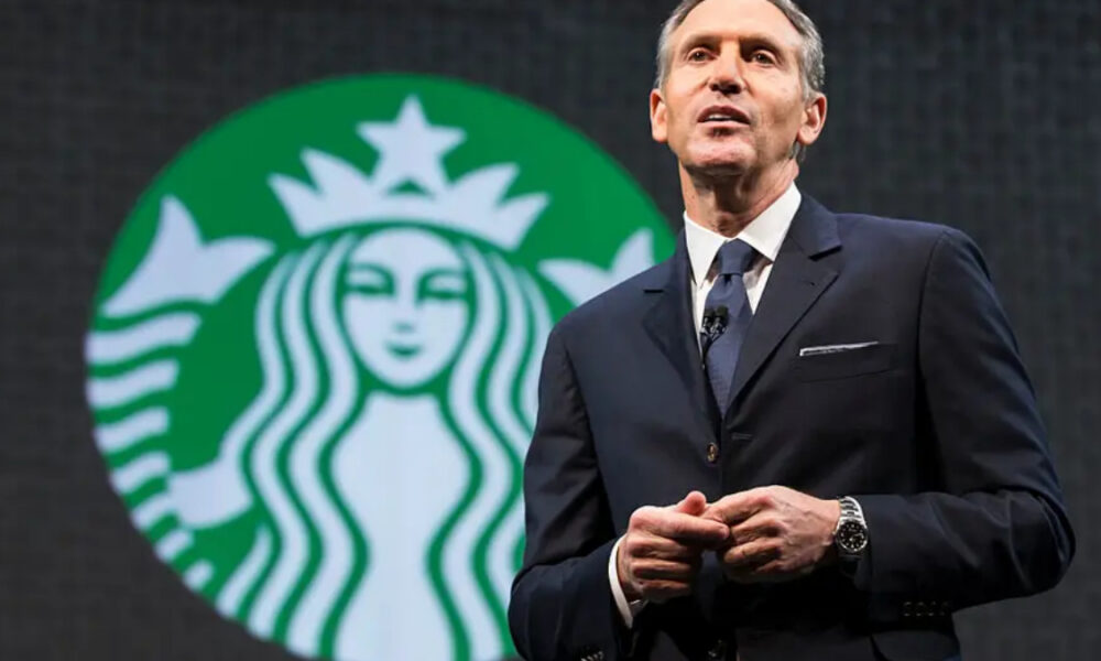 Man in a dark suit giving a speech on stage with a large Starbucks logo in the background behind him called to attention the brand scene behind him
