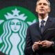 Man in a dark suit giving a speech on stage with a large Starbucks logo in the background behind him called to attention the brand scene behind him