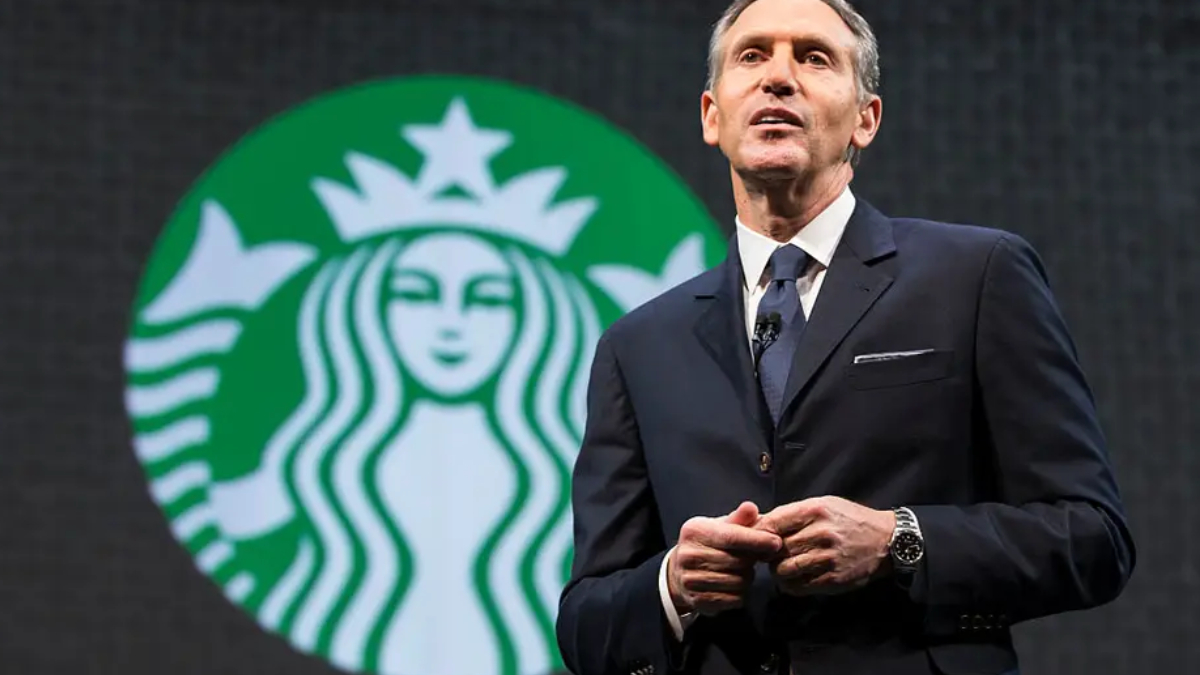 Man in a dark suit giving a speech on stage with a large Starbucks logo in the background behind him called to attention the brand scene behind him