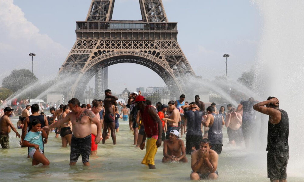 Crowd of people cooling off in a large fountain at the base of a replica Eiffel Tower, with water jets spraying around them in a sunny urban setting