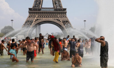 Crowd of people cooling off in a large fountain at the base of a replica Eiffel Tower, with water jets spraying around them in a sunny urban setting
