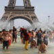 Crowd of people cooling off in a large fountain at the base of a replica Eiffel Tower, with water jets spraying around them in a sunny urban setting