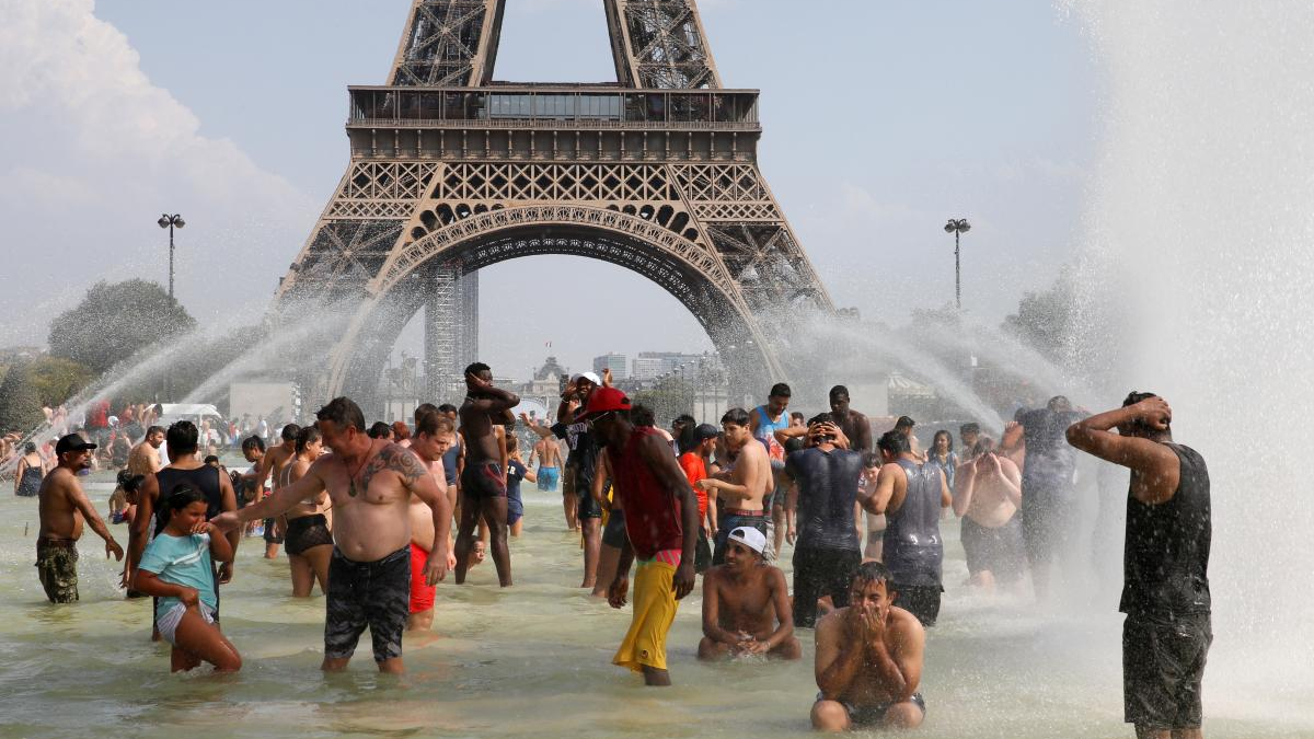 Crowd of people cooling off in a large fountain at the base of a replica Eiffel Tower, with water jets spraying around them in a sunny urban setting