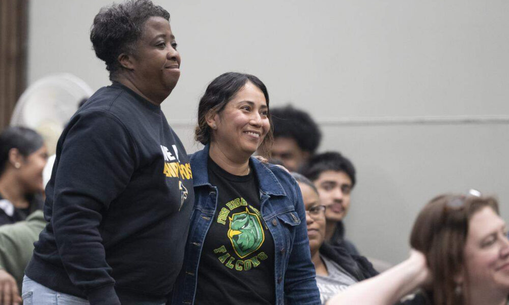 Two smiling women stand side by side at an indoor event, with seated attendees in the background.
