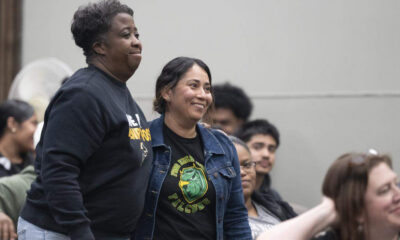 Two smiling women stand side by side at an indoor event, with seated attendees in the background.