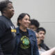 Two smiling women stand side by side at an indoor event, with seated attendees in the background.