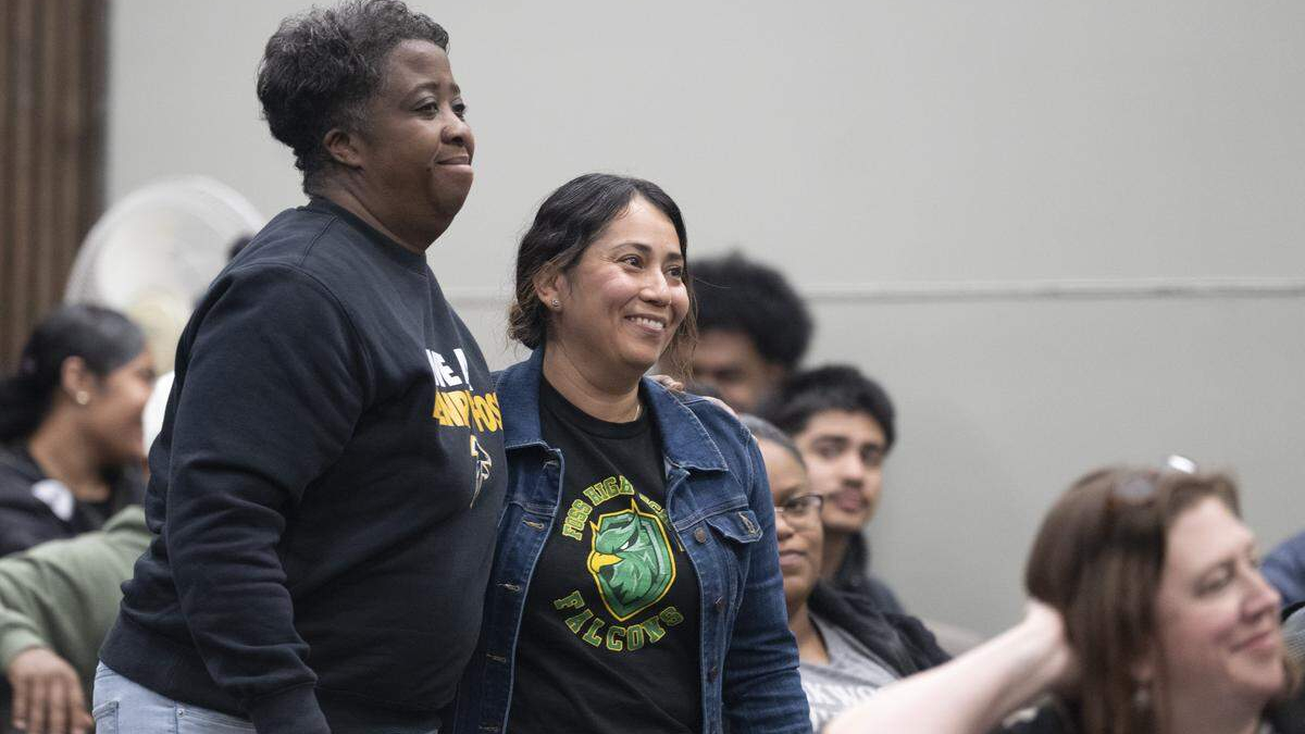 Two smiling women stand side by side at an indoor event, with seated attendees in the background.