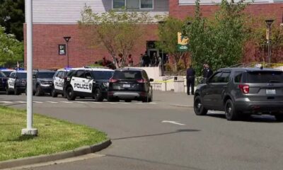 Police cars block a street outside a brick building with yellow tape and onlookers nearby.