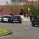 Police cars block a street outside a brick building with yellow tape and onlookers nearby.