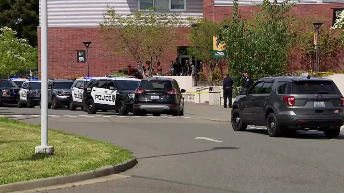 Police cars block a street outside a brick building with yellow tape and onlookers nearby.