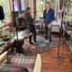 Two people converse in a cozy home studio filled with musical instruments and recording gear.