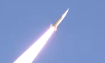 Rocket launch: a bright exhaust plume trails upward through a clear blue sky as a rocket ascends.