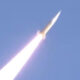 Rocket launch: a bright exhaust plume trails upward through a clear blue sky as a rocket ascends.