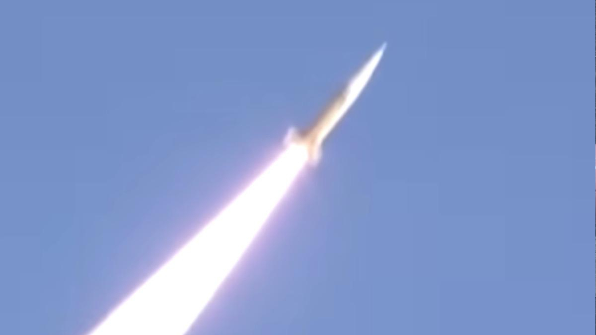 Rocket launch: a bright exhaust plume trails upward through a clear blue sky as a rocket ascends.