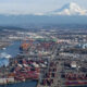 Busy cargo port with colorful containers and blue cranes along the waterfront, mountain in the background.