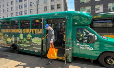 Green King County Metro waterfront shuttle bus with doors open, a man carrying an orange bag stepping onto the sidewalk.