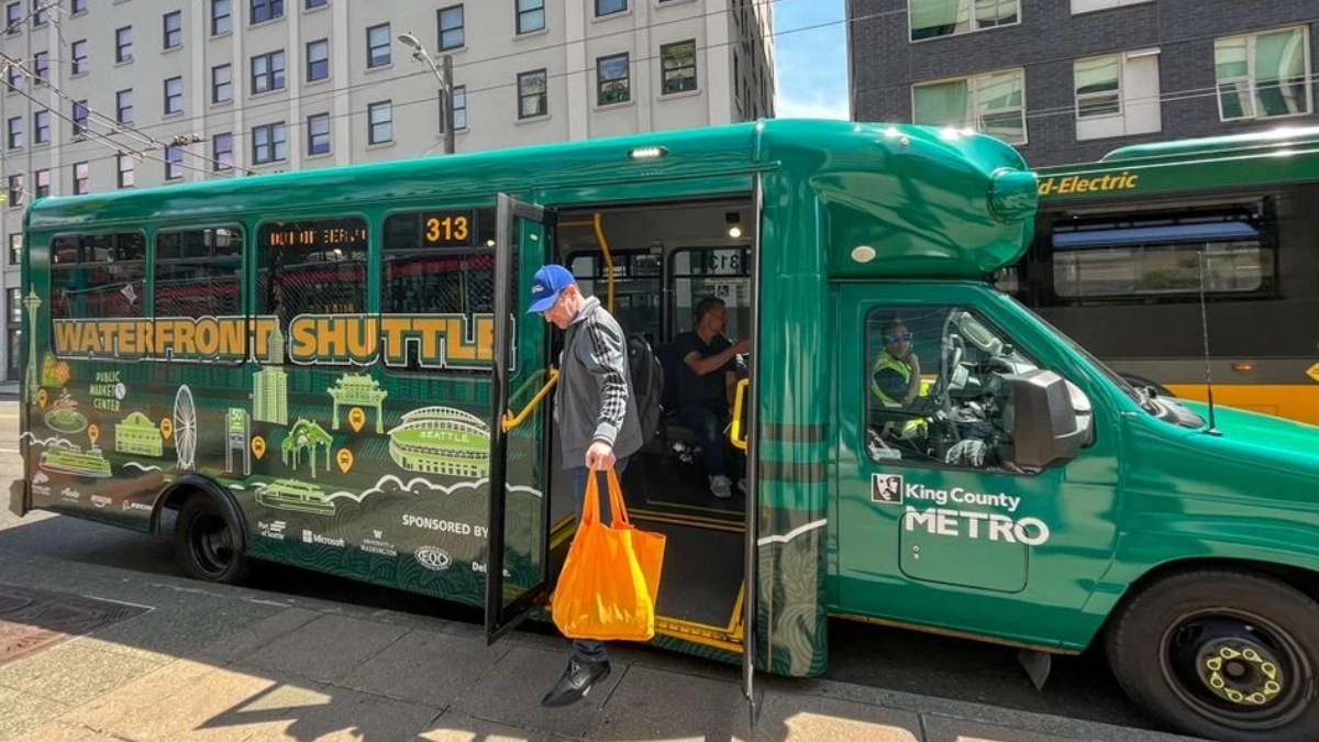 Green King County Metro waterfront shuttle bus with doors open, a man carrying an orange bag stepping onto the sidewalk.