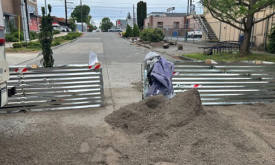 A street blocked by metal barriers with a dirt pile in the foreground and a purple tarp nearby, indicating construction.
