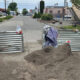 A street blocked by metal barriers with a dirt pile in the foreground and a purple tarp nearby, indicating construction.