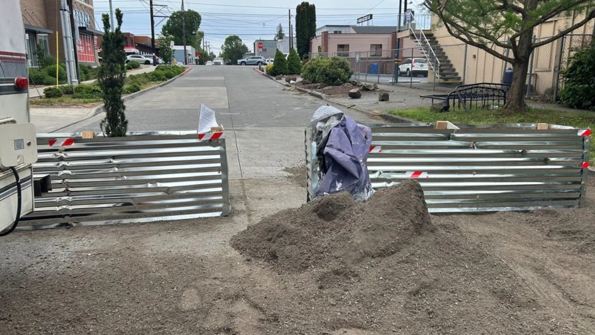 A street blocked by metal barriers with a dirt pile in the foreground and a purple tarp nearby, indicating construction.
